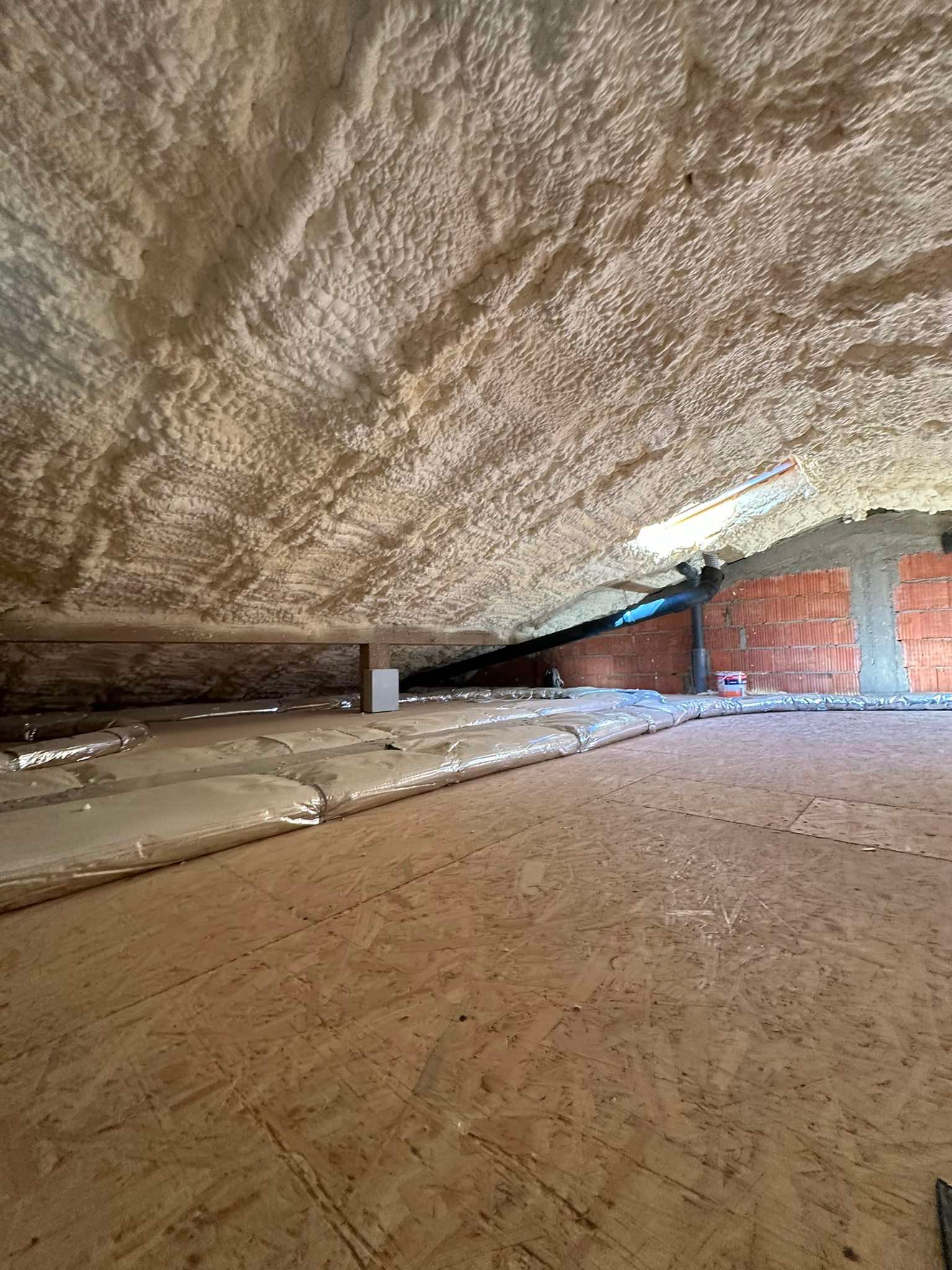 Poddasze po ociepleniu pianką PUR, widoczna struktura pianki na skosach dachu, podłoga wyłożona płytami OSB, fragment instalacji wentylacyjnej i fragment ściany z czerwonej cegły.