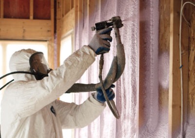 Pracownik w kombinezonie ochronnym natryskuje pianę izolacyjną na ściany szkieletowe budynku, widoczne drewniane elementy konstrukcyjne.