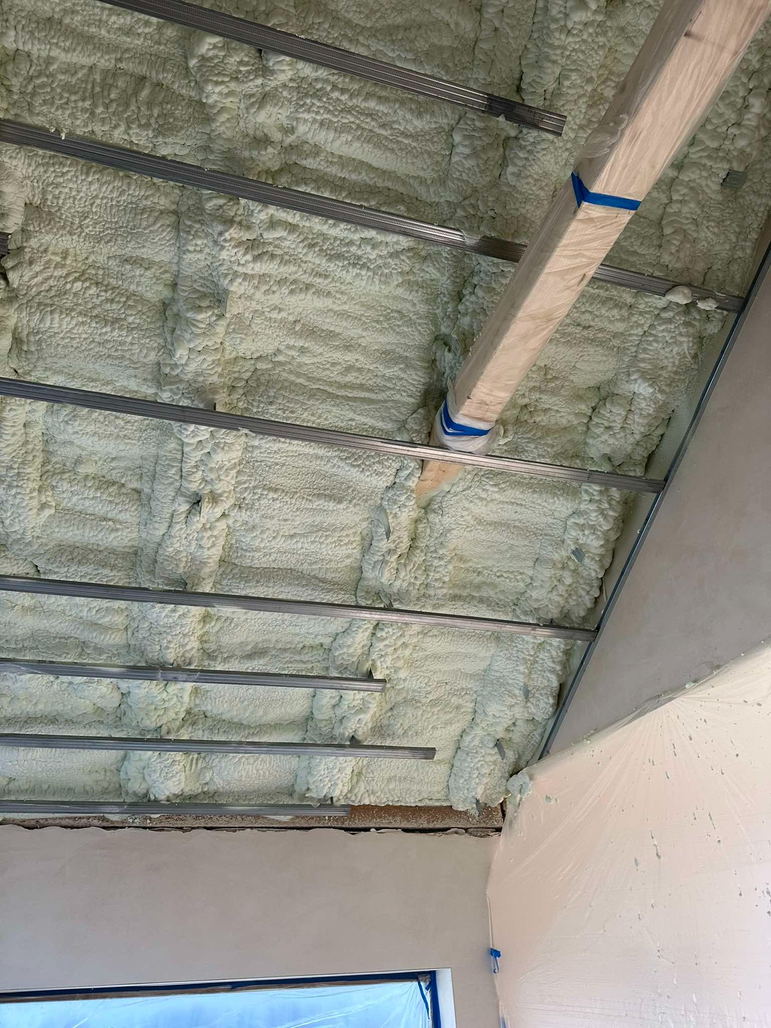 Świeżo nałożona, jasna pianka izolacyjna między metalowymi profilami sufitowymi, zabezpieczona folią malarską na ścianie, widoczny fragment okna.