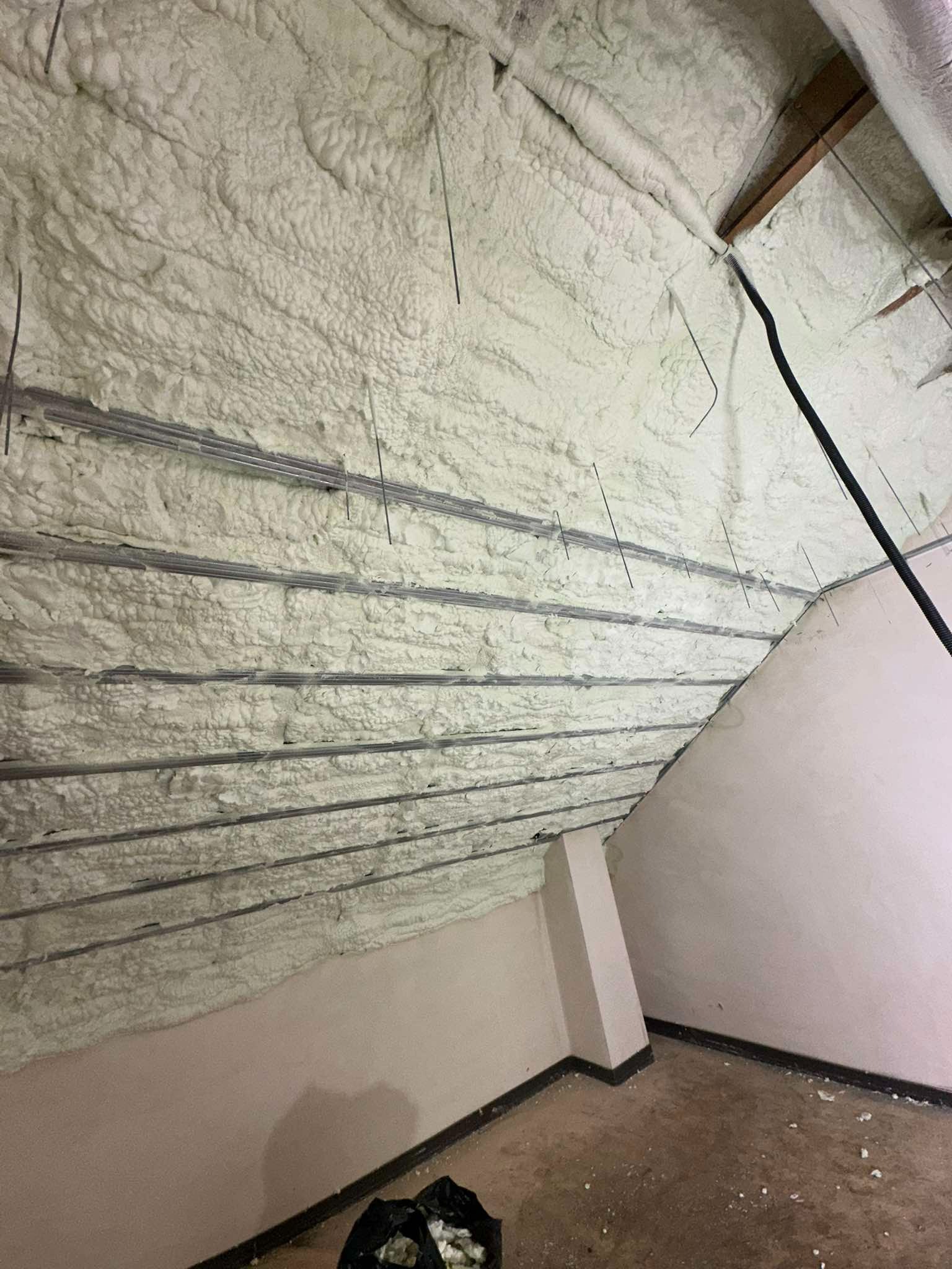 Poddasze po ociepleniu pianką PUR, widoczne metalowe elementy konstrukcyjne wystające z piany, ściana boczna pomalowana na jasny kolor, na podłodze resztki materiałów budowlanych w czarnym worku.