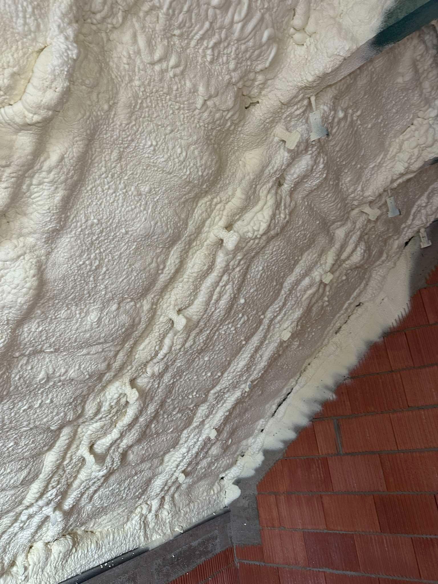 Świeżo nałożona, jasna pianka izolacyjna na poddaszu, widoczne mocowania i fragment muru z czerwonej cegły.