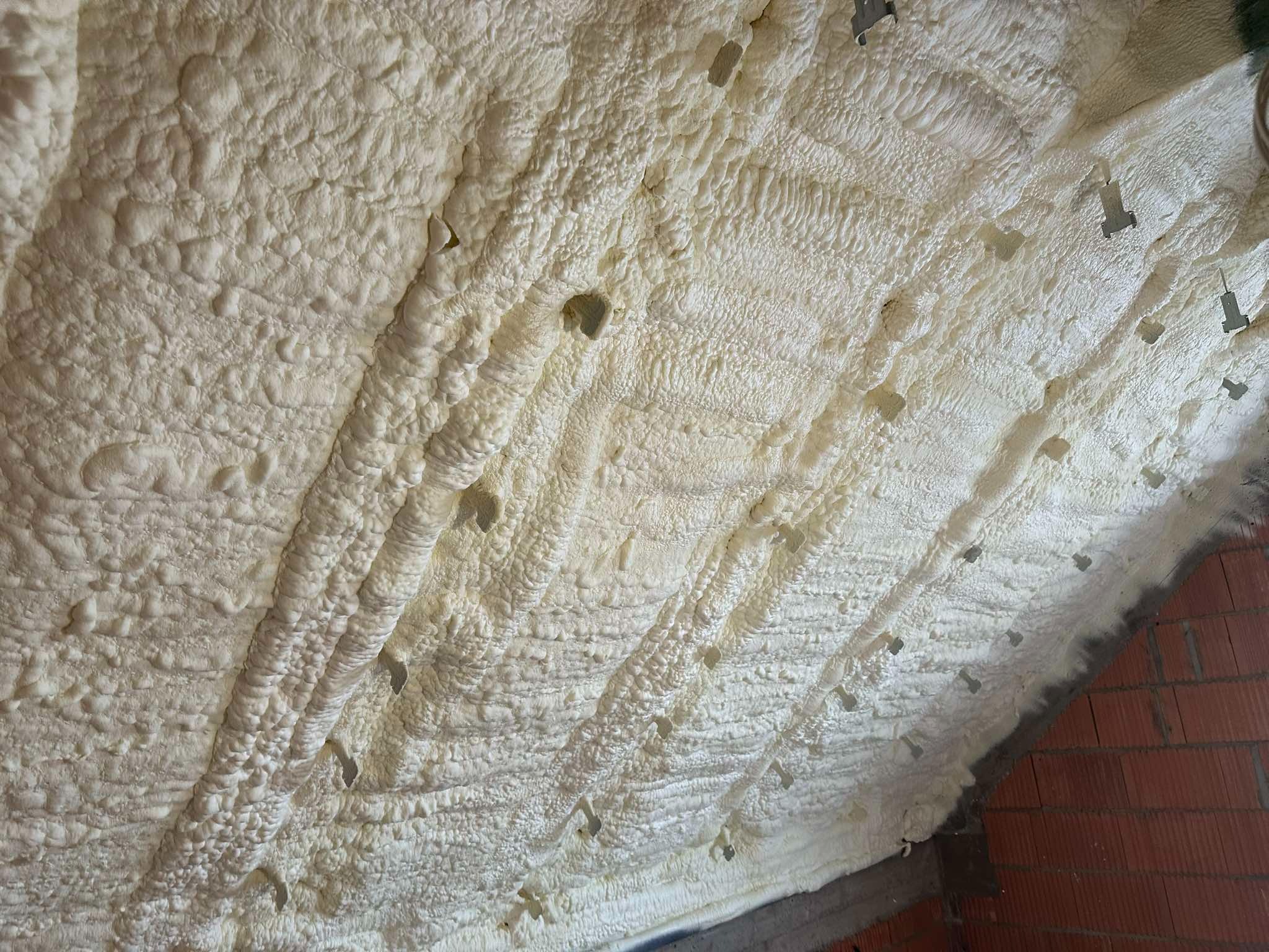 Świeżo nałożona, biała pianka izolacyjna PUR na poddaszu w Bełku, widoczne metalowe uchwyty mocujące i fragment ściany z cegły.