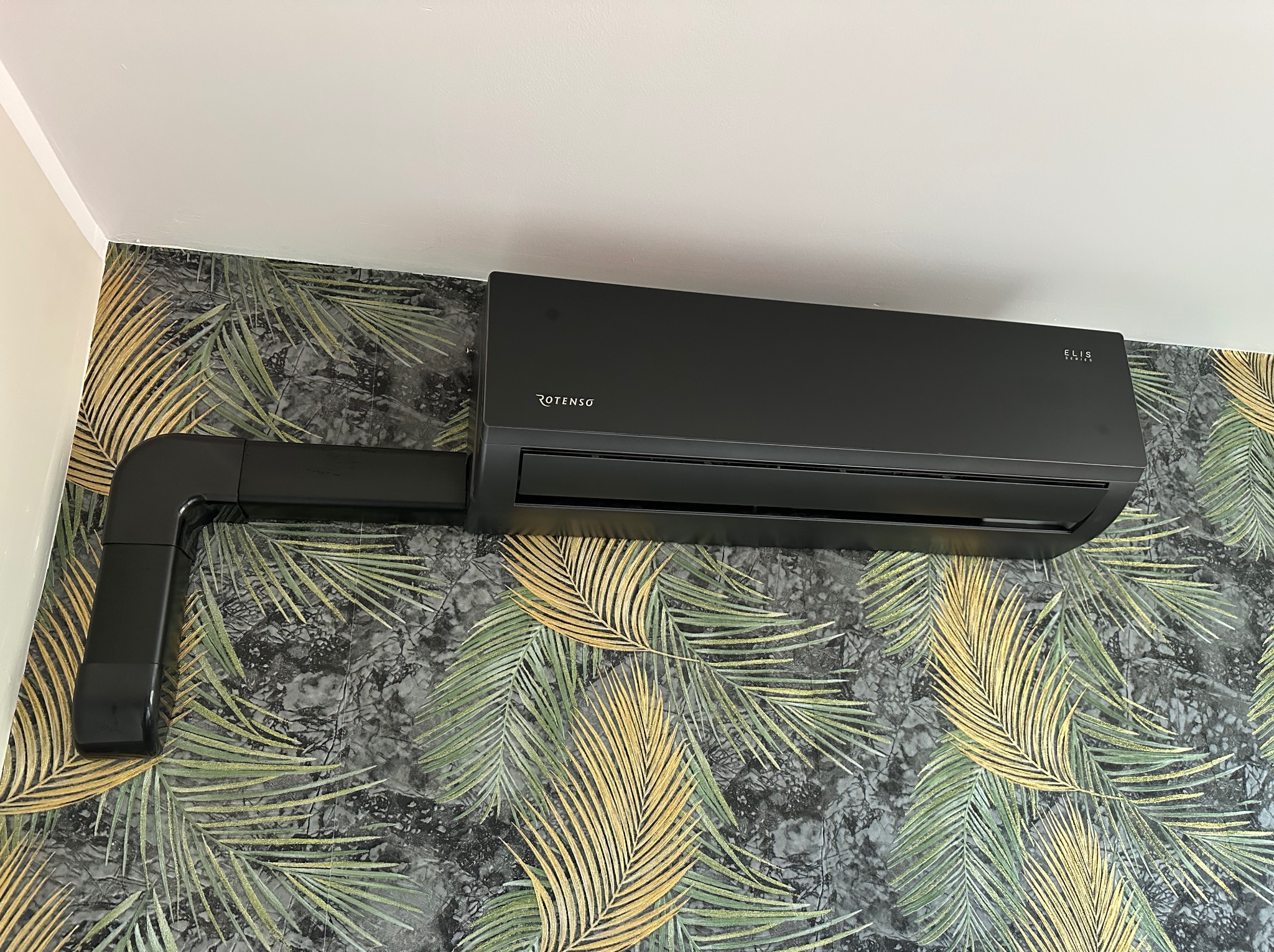 Czarna jednostka klimatyzacji Rotenso Elis Series zamontowana na ścianie z tapetą w motywy liści palmowych.