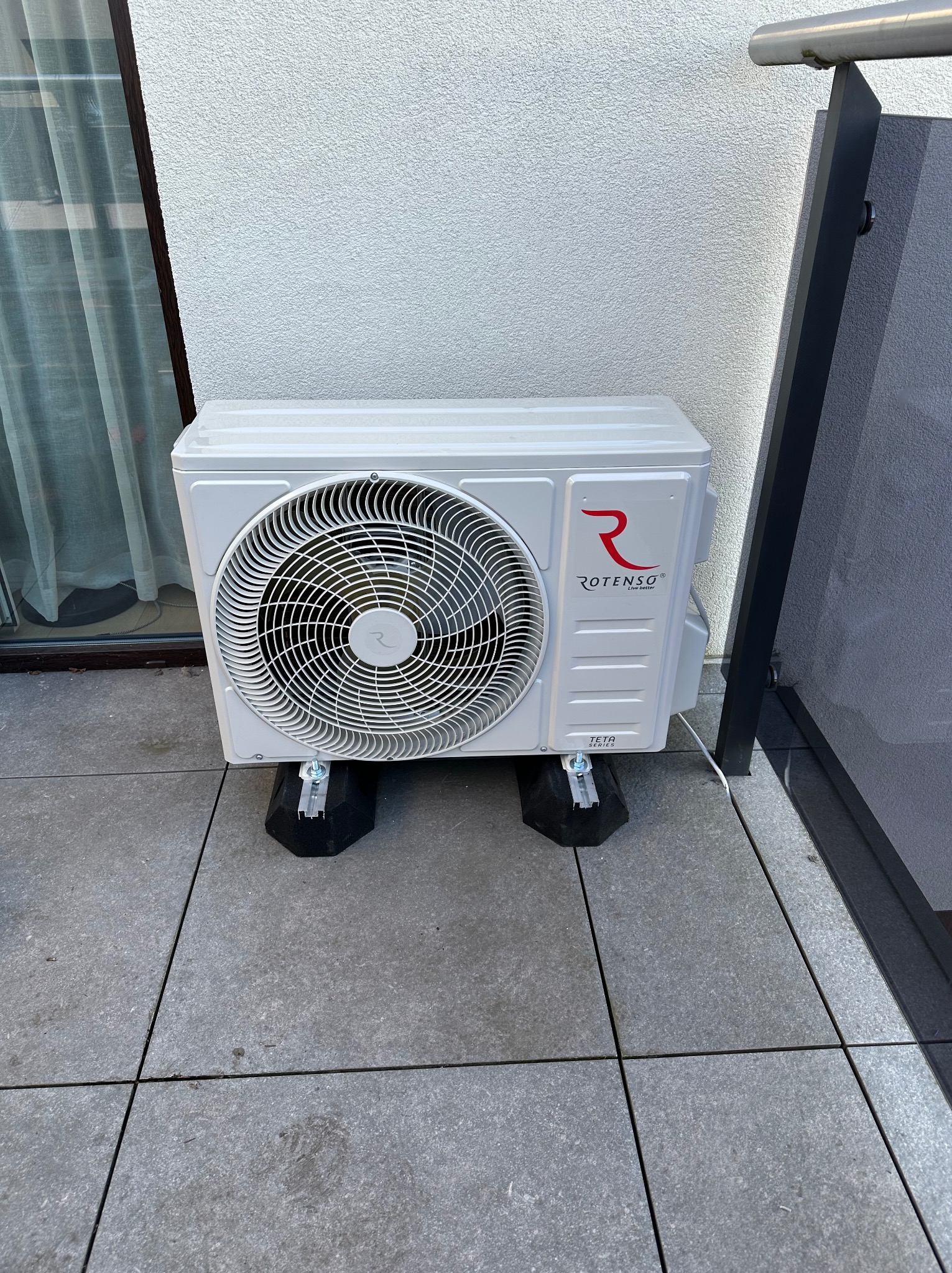 Zewnętrzna jednostka klimatyzacji Rotenso Teta Series, umieszczona na czarnych podkładkach antywibracyjnych na szarym tarasie, z widoczną ścianą budynku i fragmentem balustrady.