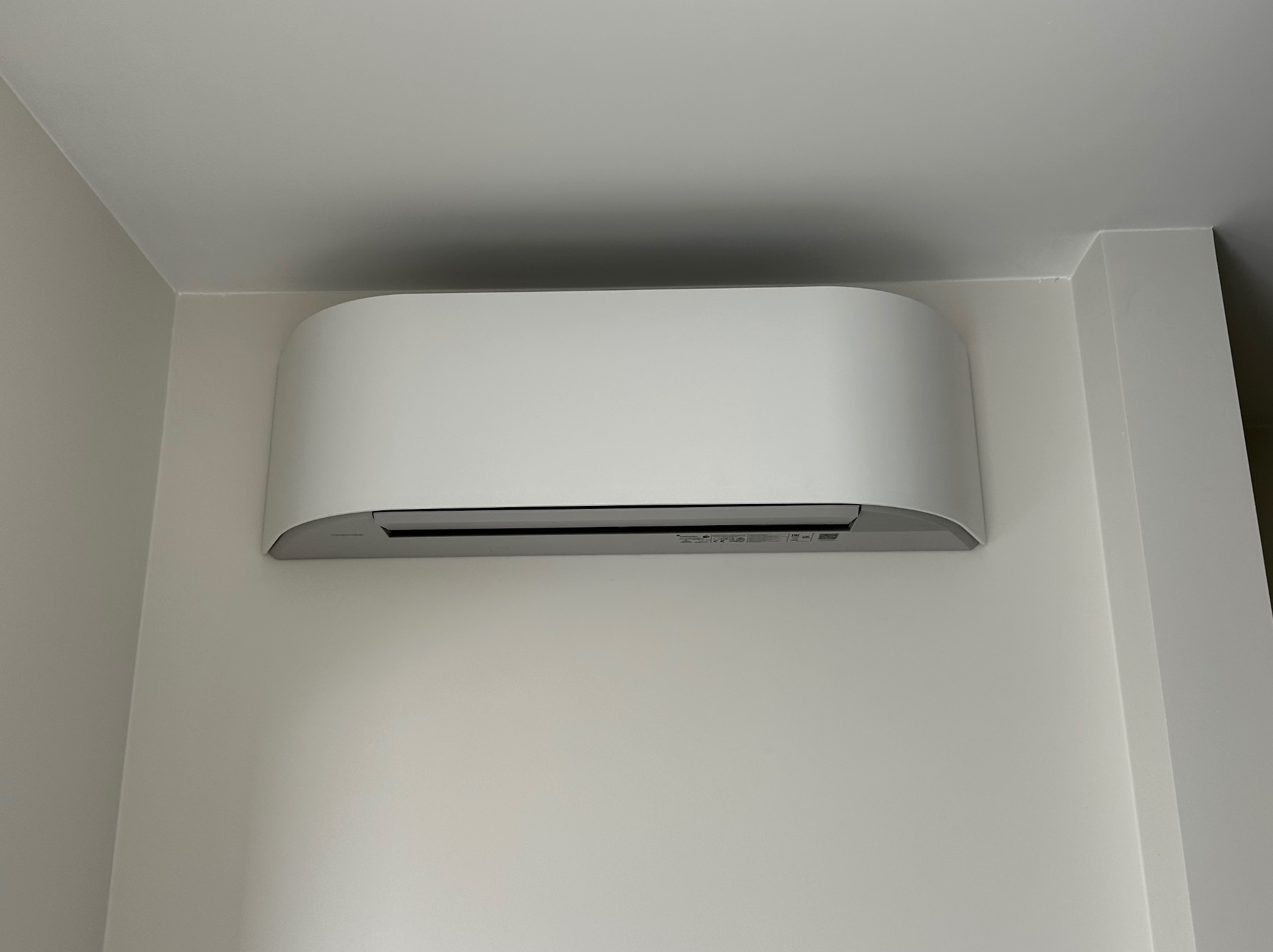 Nowoczesny, biały klimatyzator ścienny zamontowany w rogu jasnego pomieszczenia, widoczne delikatne cienie na ścianie.