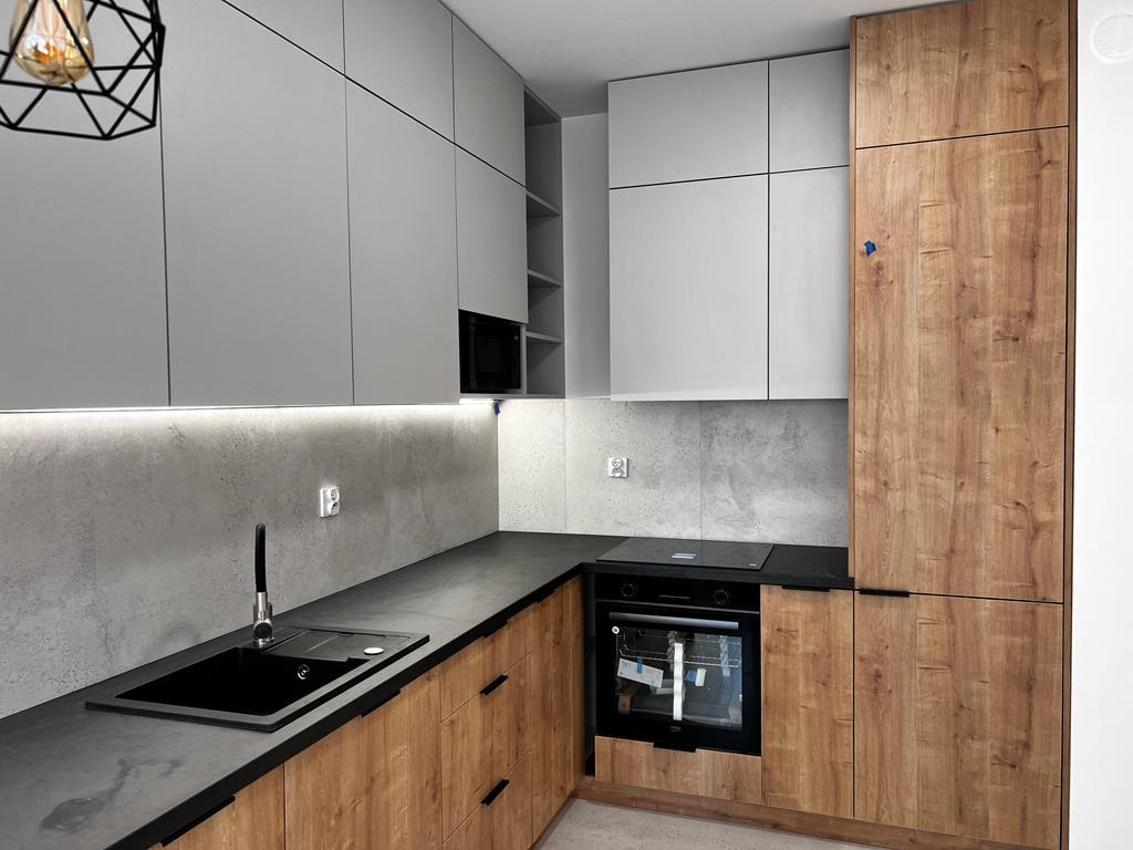 Nowoczesna kuchnia z szarymi górnymi szafkami i drewnopodobnymi dolnymi szafkami, czarnym blatem i zlewozmywakiem, oświetleniem LED pod szafkami górnymi oraz wiszącą lampą geometryczną.