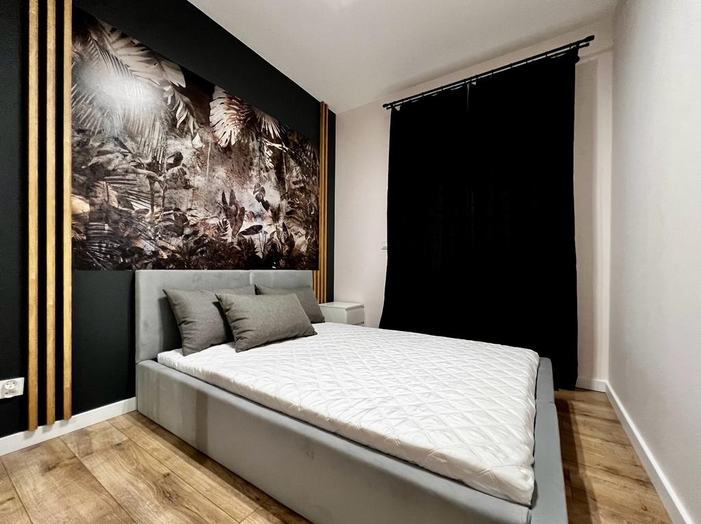 Sypialnia z łóżkiem w szarej ramie, białym pikowanym materacem i szarymi poduszkami, ściana z fototapetą w motywy roślinne w stylu dżungli, drewniane lamele na ścianie i czarne zasłony w oknie.
