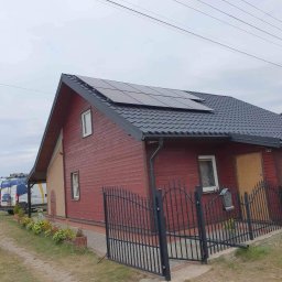 Perfect Instal Michał Wszeborowski - Czerwony dom jednorodzinny z czarnym dachem i panelami słonecznymi, widok z zewnątrz, ogrodzenie z czarnego metalu, trawnik i zaparkowany biały bus z logo firmy w tle.