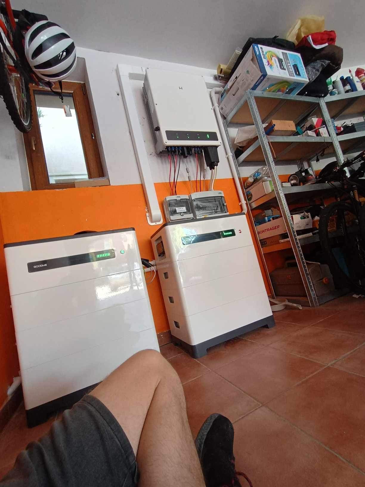 Dwa białe magazyny energii GoodWe w garażu z pomarańczowymi ścianami, inwerter zamontowany na ścianie, widoczny fragment nogi i buta osoby robiącej zdjęcie.