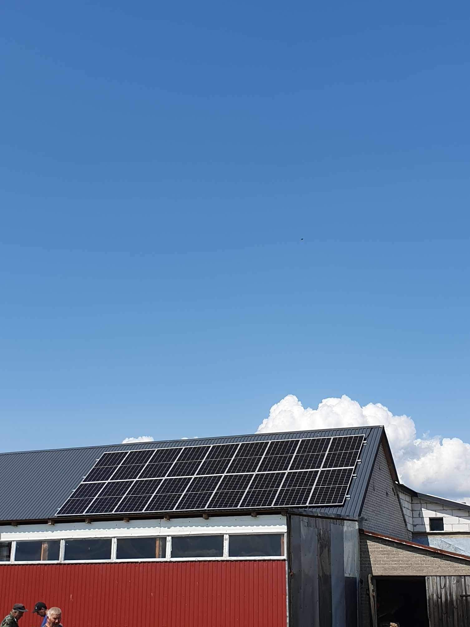 Panele słoneczne na dachu budynku gospodarczego z czerwonymi ścianami i szarym dachem, na tle błękitnego nieba z białymi obłokami.