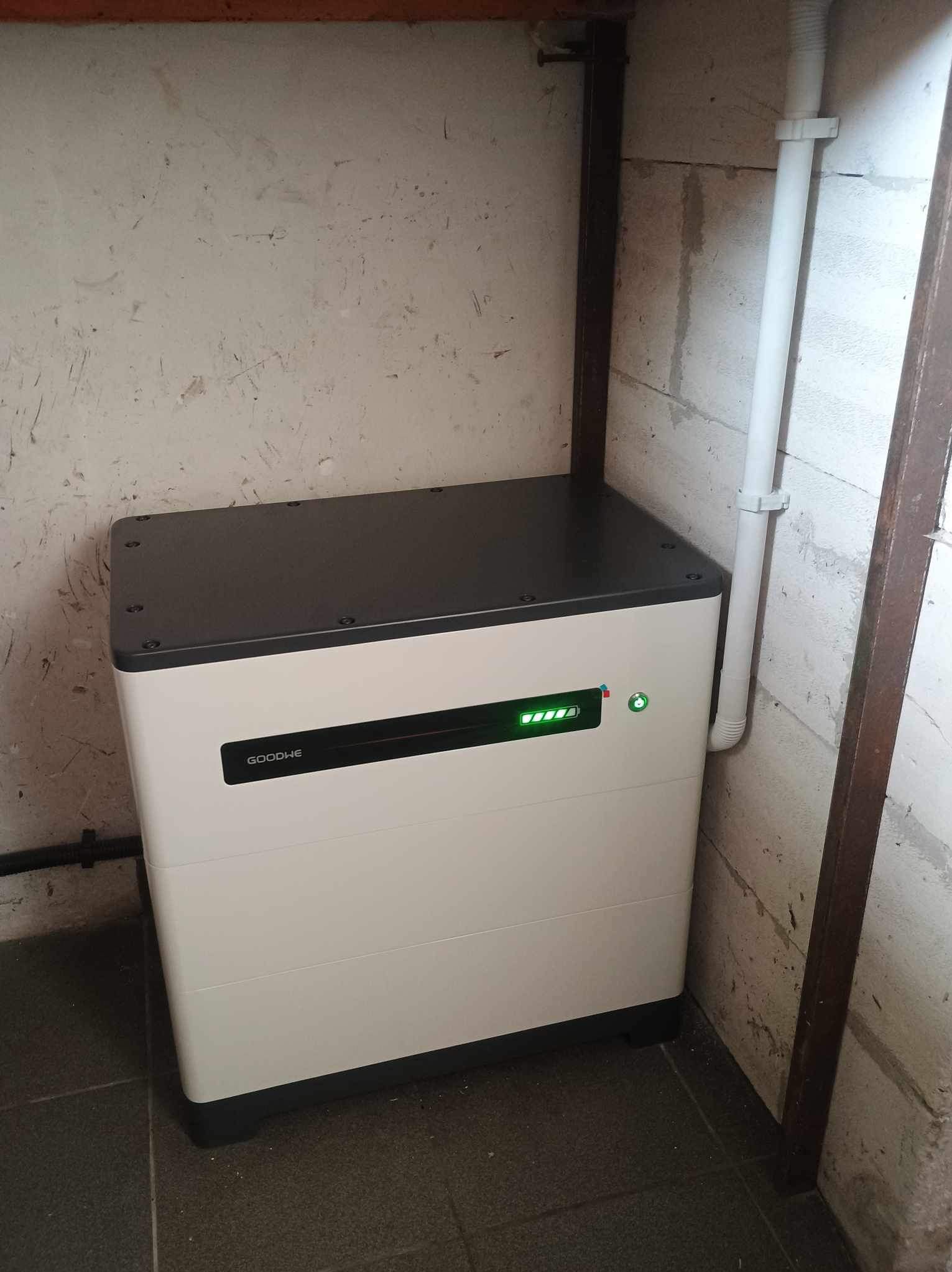 Biały magazyn energii GoodWe z czarnym blatem, umieszczony w rogu pomieszczenia o surowych, betonowych ścianach i ciemnej podłodze, z widocznymi wskaźnikami naładowania.