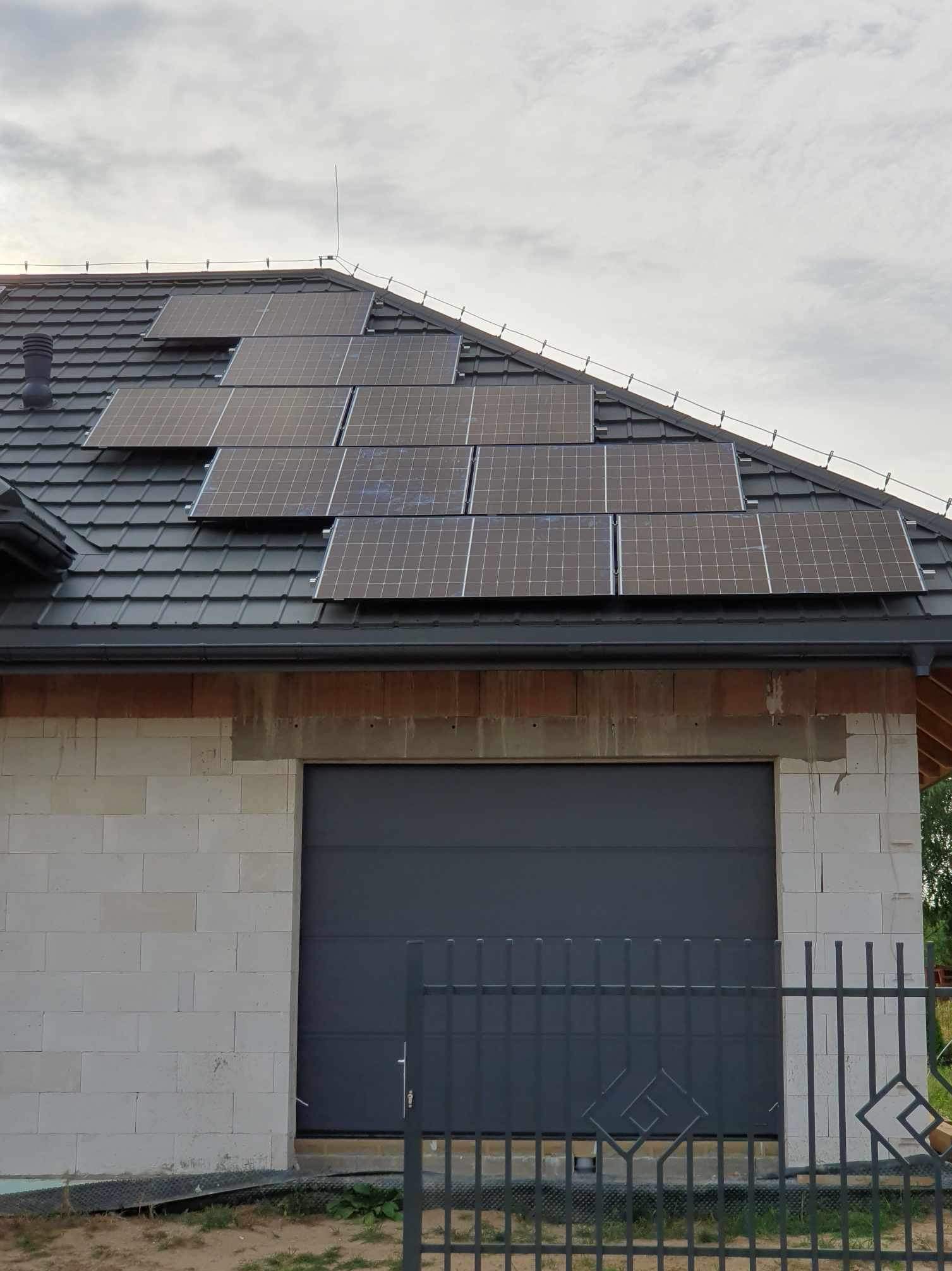 Panele słoneczne na dachu domu z garażem, widok z dołu, ogrodzenie w tle. Dachówka ceramiczna w kolorze grafitowym.