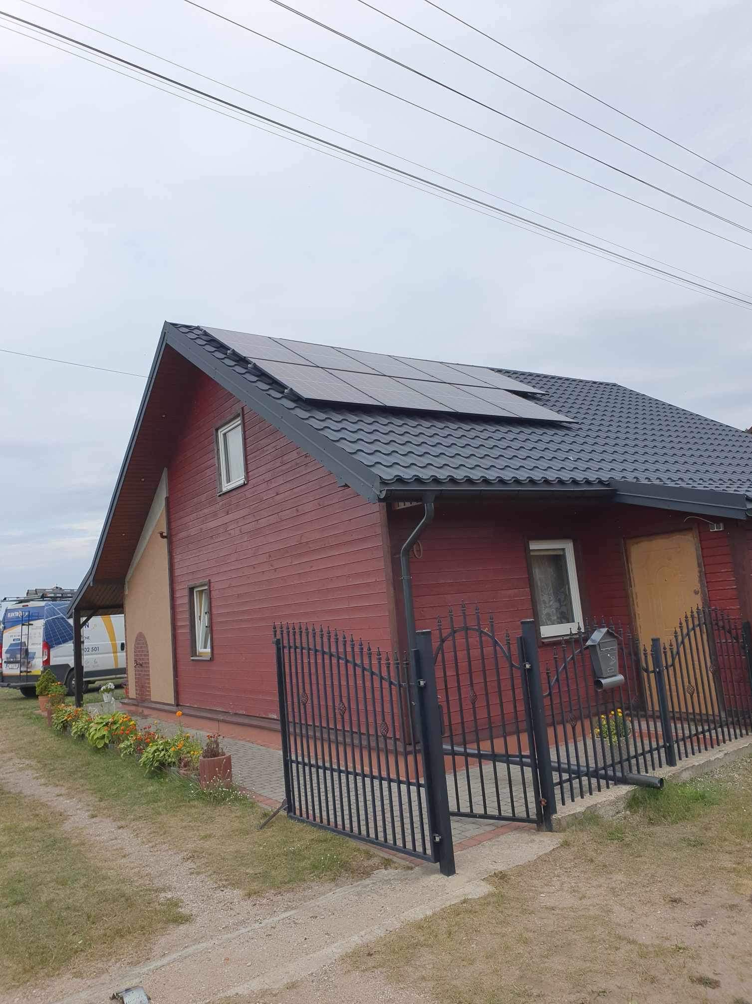 Czerwony dom jednorodzinny z czarnym dachem i panelami słonecznymi, widok z zewnątrz, ogrodzenie z czarnego metalu, trawnik i zaparkowany biały bus z logo firmy w tle.