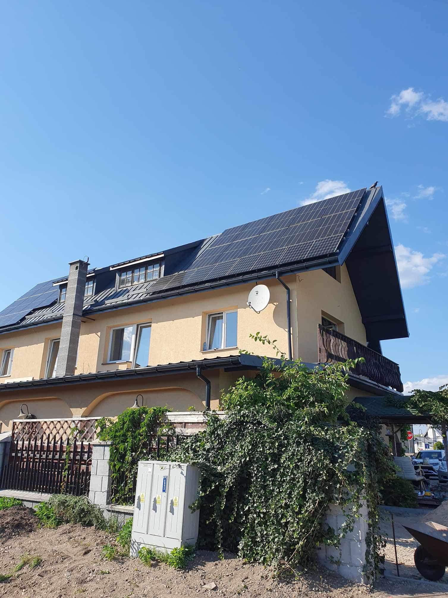 Dom jednorodzinny z żółtą elewacją i ciemnymi panelami słonecznymi na dachu, z widoczną instalacją satelitarną na ścianie szczytowej oraz skrzynką elektryczną przy ogrodzeniu.