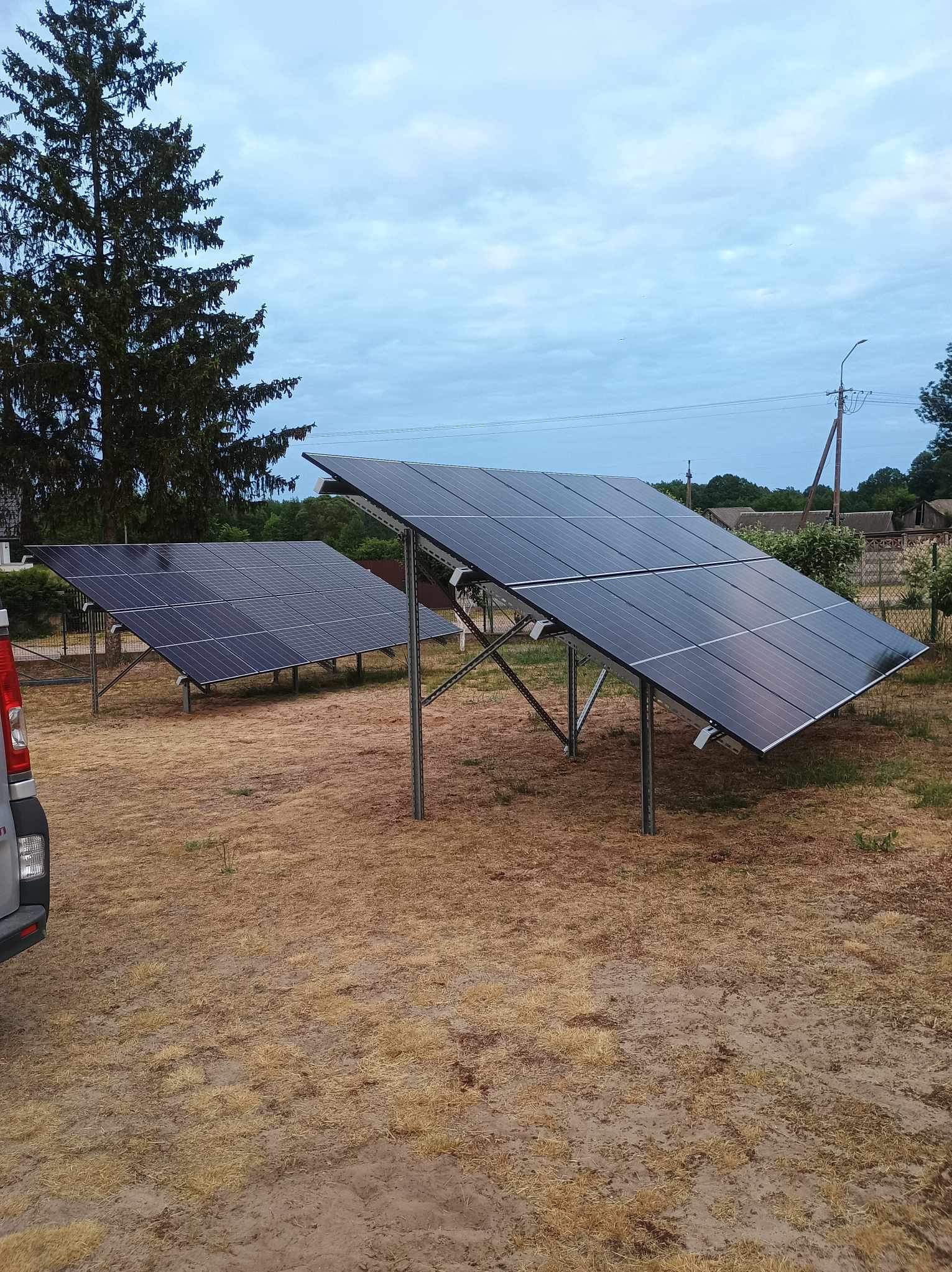 Dwa rzędy paneli słonecznych na metalowych stelażach, zamontowane na wyschniętej trawie, z widocznym fragmentem samochodu i drzewem w tle.
