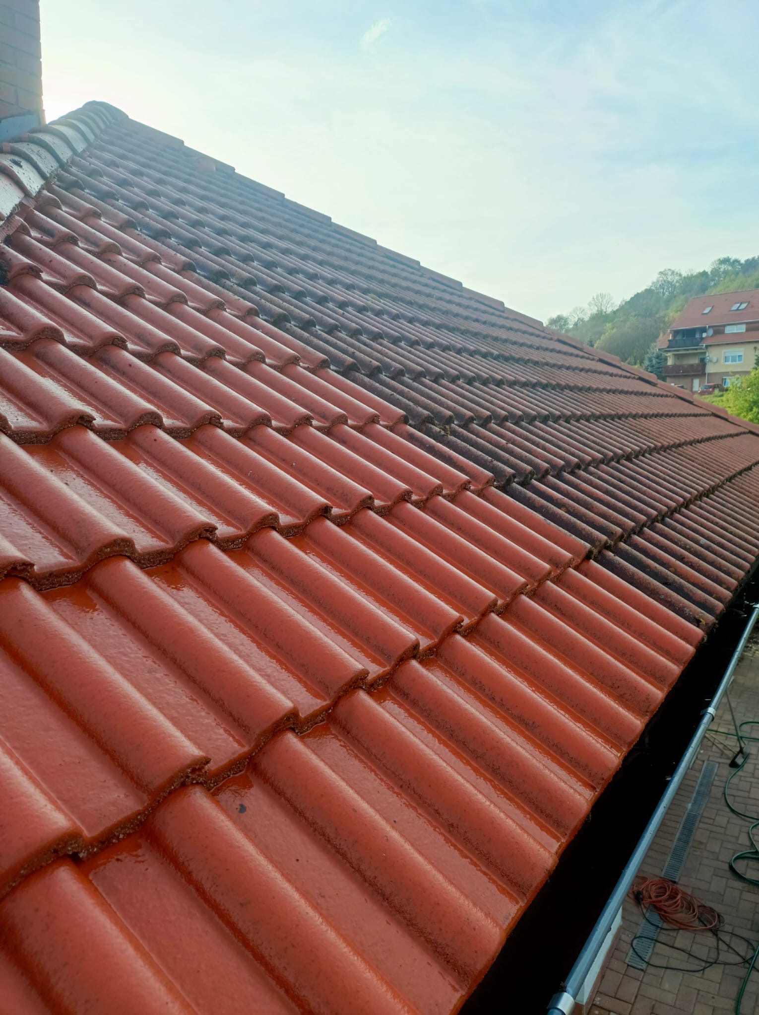 Częściowo umyty dach z czerwonej dachówki, widoczna różnica w czystości między umytą a nieumytą powierzchnią, zarys budynku w tle.