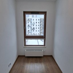 ZAMALOWANI - Pusty pokój po remoncie: brązowe panele podłogowe, białe ściany, okno z widokiem na blok i ozdobny żyrandol. Minimalistyczne wnętrze po odświeżeniu.