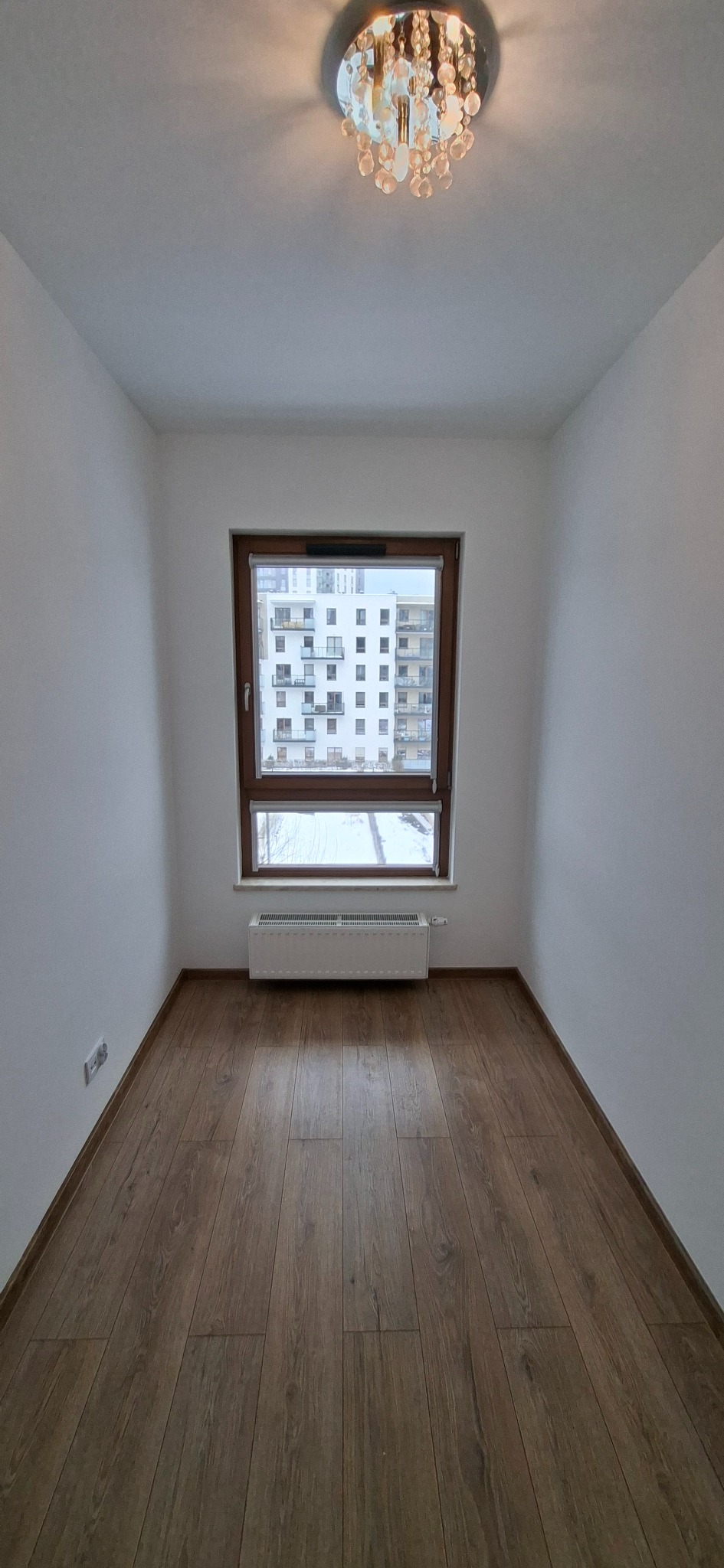Pusty pokój po remoncie: brązowe panele podłogowe, białe ściany, okno z widokiem na blok i ozdobny żyrandol. Minimalistyczne wnętrze po odświeżeniu.