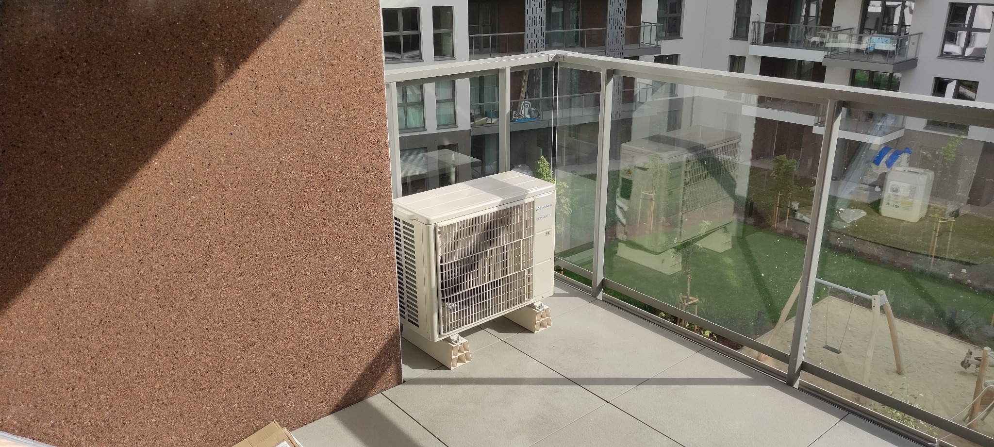 Zewnętrzna jednostka klimatyzacji zamontowana na balkonie nowoczesnego apartamentowca, widok na plac zabaw przez szklaną balustradę.