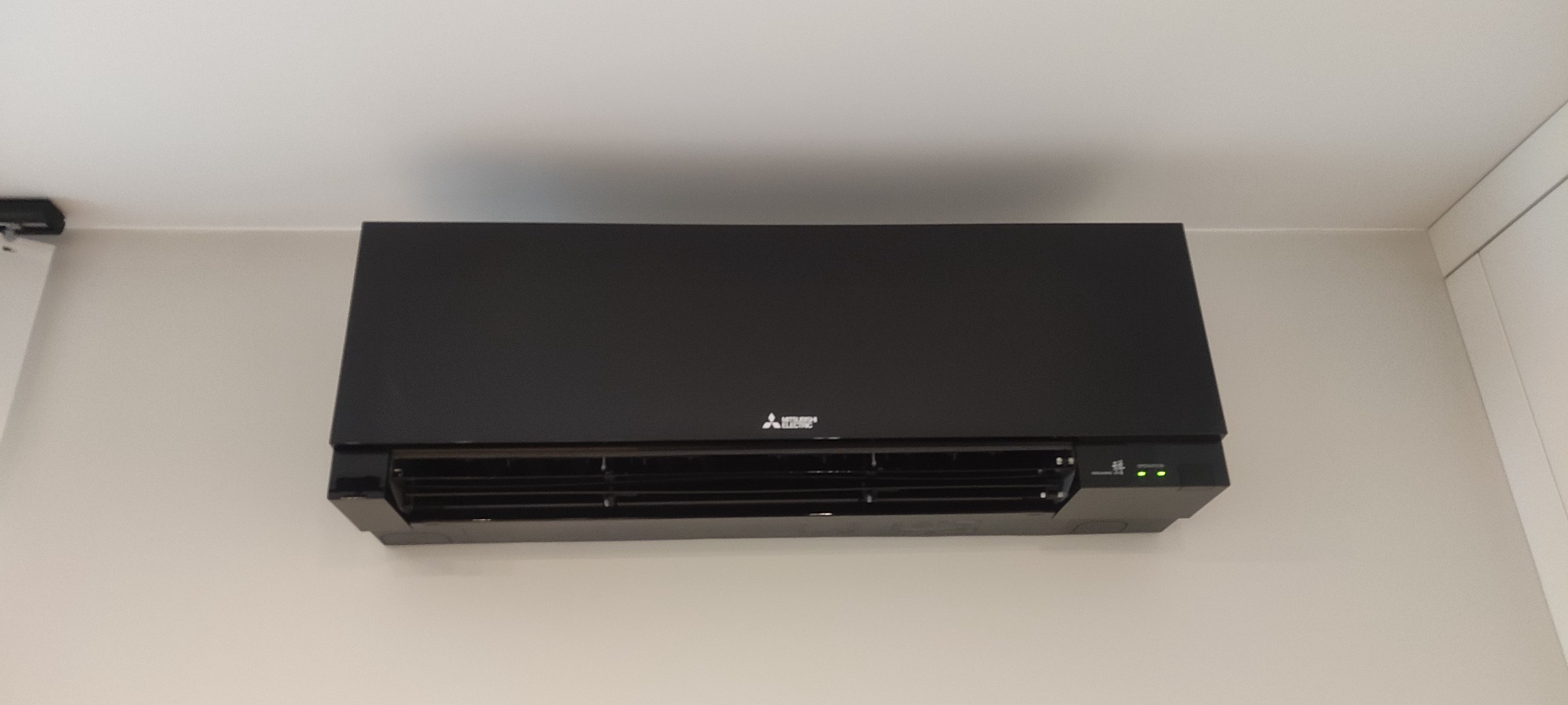 Czarna jednostka klimatyzacji Mitsubishi Electric zamontowana na ścianie w pomieszczeniu o jasnych ścianach, widoczne diody sygnalizacyjne.