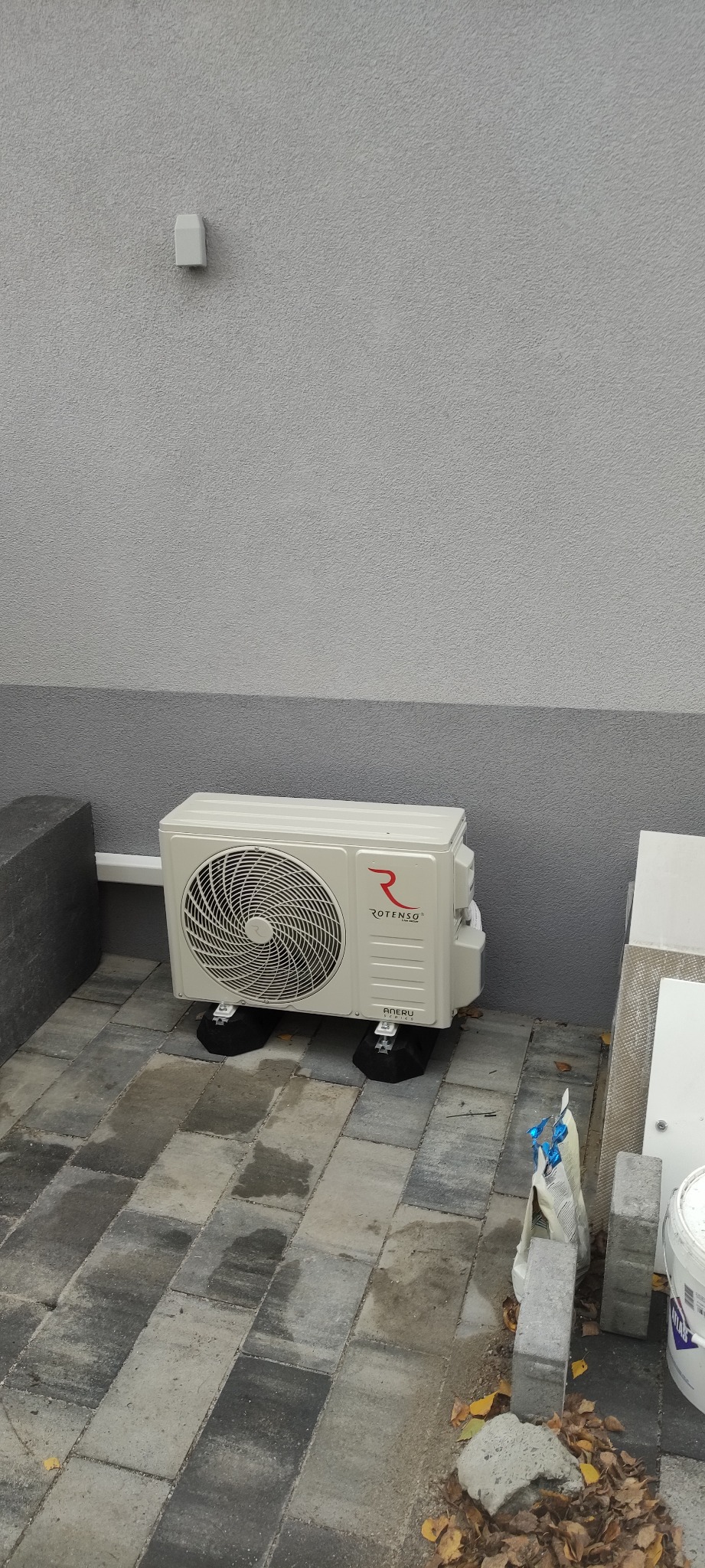 Zewnętrzna jednostka klimatyzacji Rotenso na czarnych podkładkach antywibracyjnych, umieszczona na szarej kostce brukowej przy elewacji budynku o nierównej fakturze.