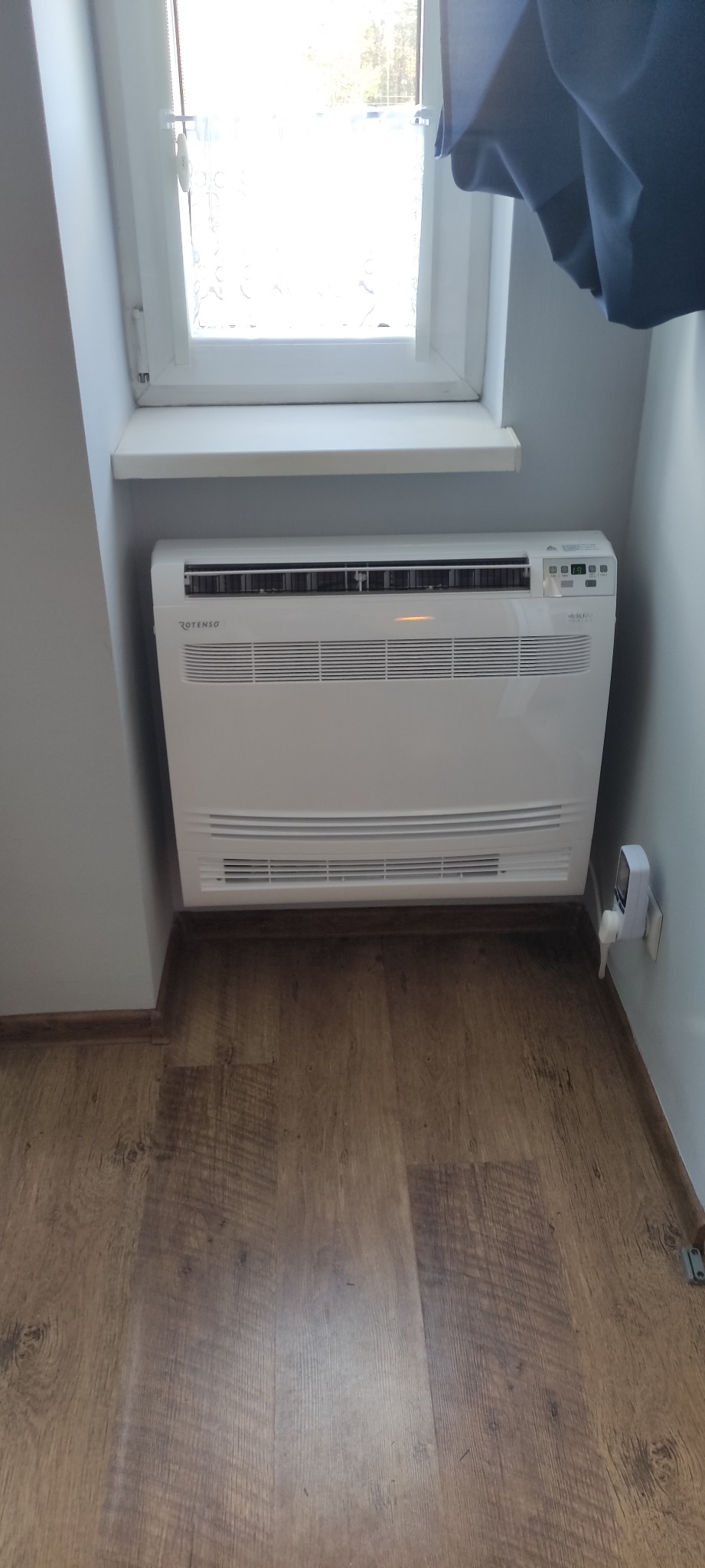 Biały klimatyzator pod oknem na poddaszu, widoczny brązowy panel podłogowy i fragment niebieskiej zasłony.