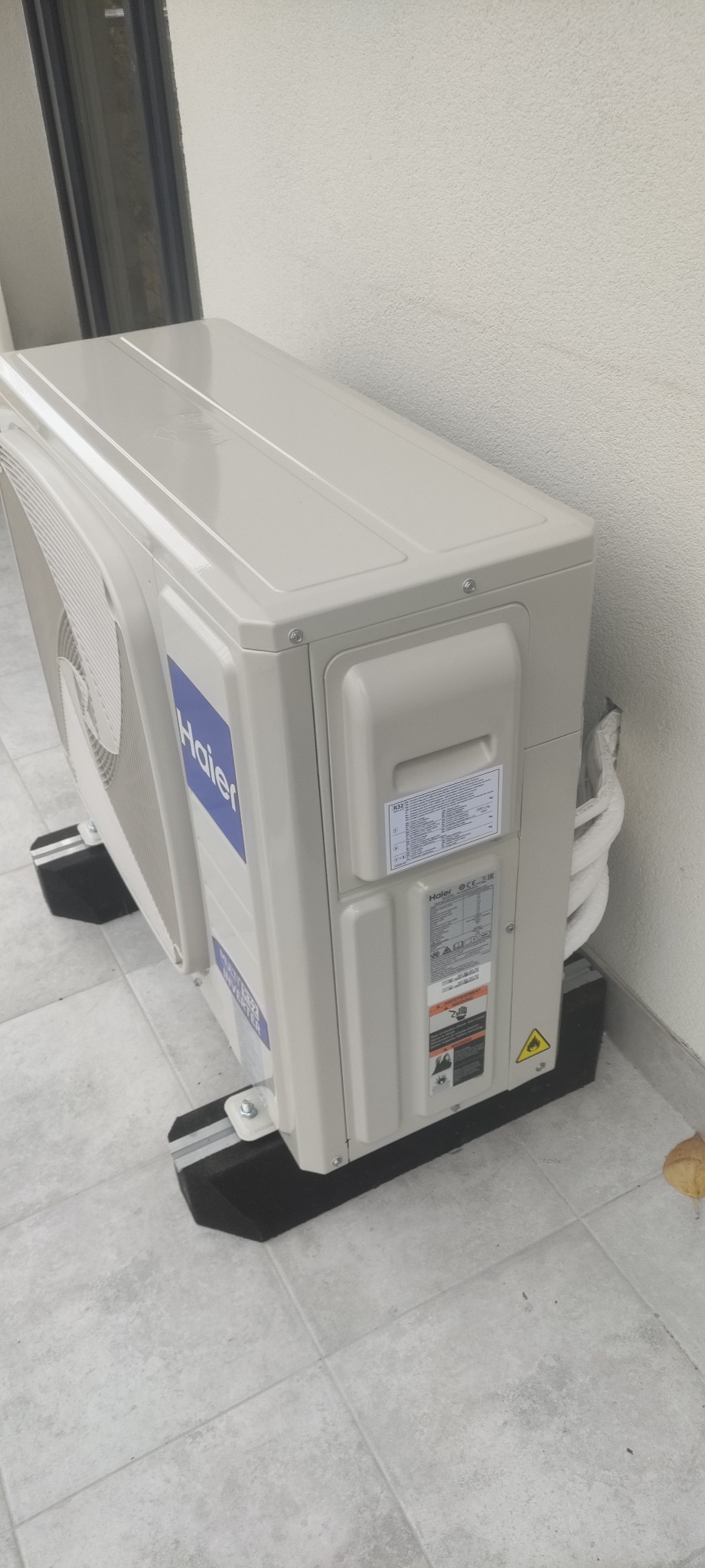 Zewnętrzna jednostka klimatyzacji Haier zamontowana na wspornikach na płytkach, widoczne przewody chłodnicze i naklejki informacyjne.