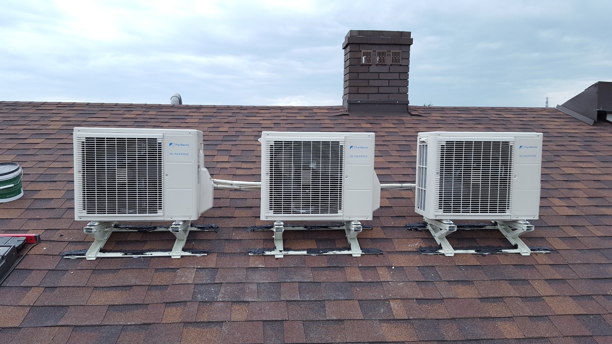 Trzy identyczne jednostki zewnętrzne klimatyzacji Fuji Electric DC Inverter zamontowane na wspornikach na brązowym dachu z gontów bitumicznych, widoczny komin wentylacyjny.