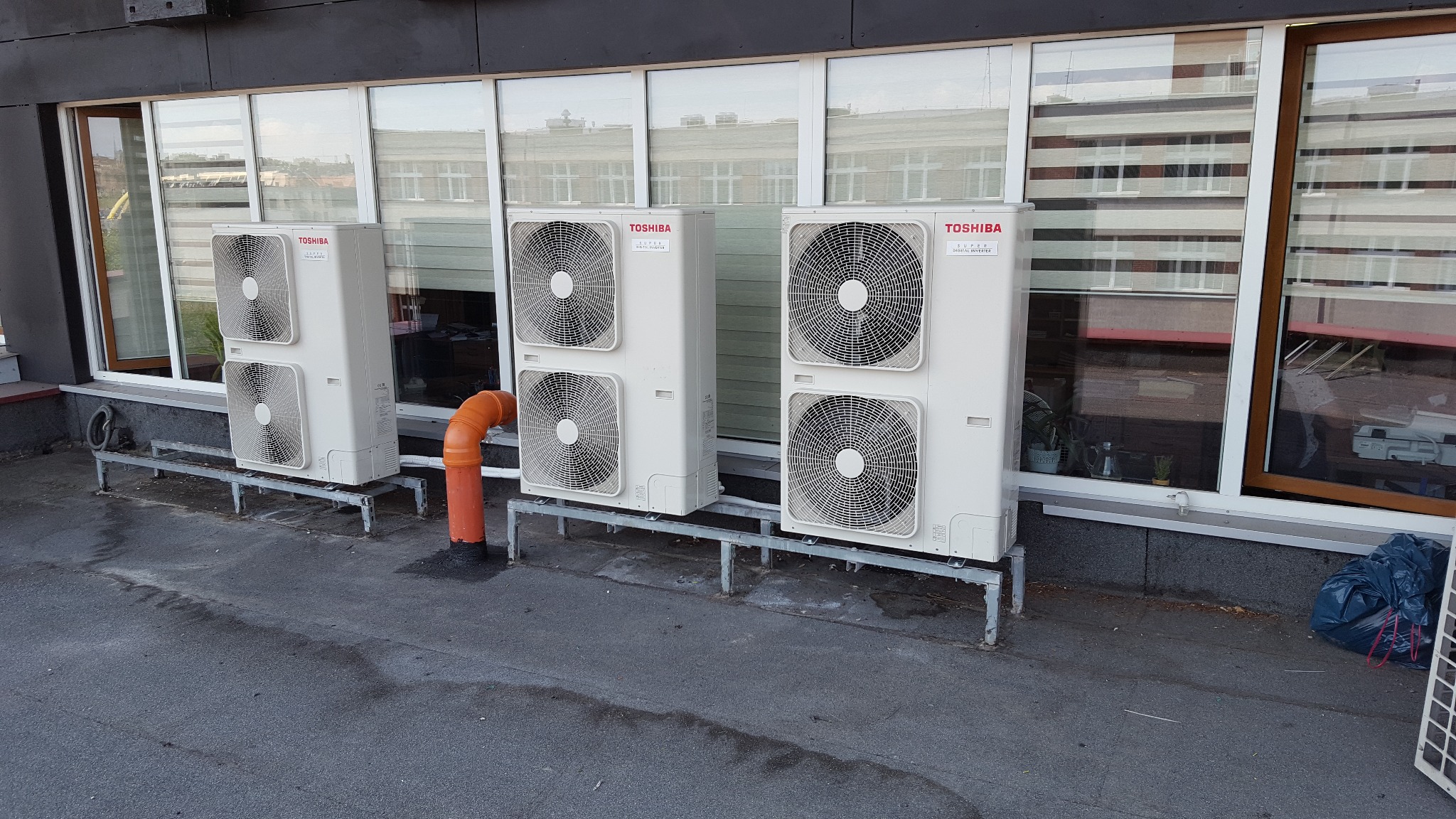 Trzy zewnętrzne jednostki klimatyzacji Toshiba zamontowane na dachu budynku komercyjnego z pomarańczową rurą odprowadzającą skropliny.