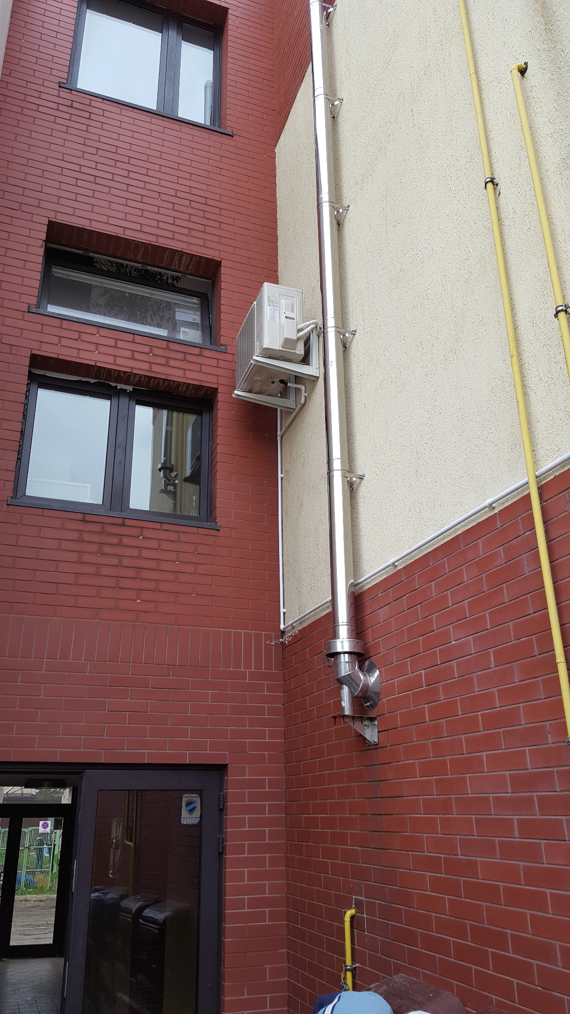 Zewnętrzna jednostka klimatyzacji z odprowadzeniem skroplin po elewacji budynku, widoczne rury gazowe i okna.