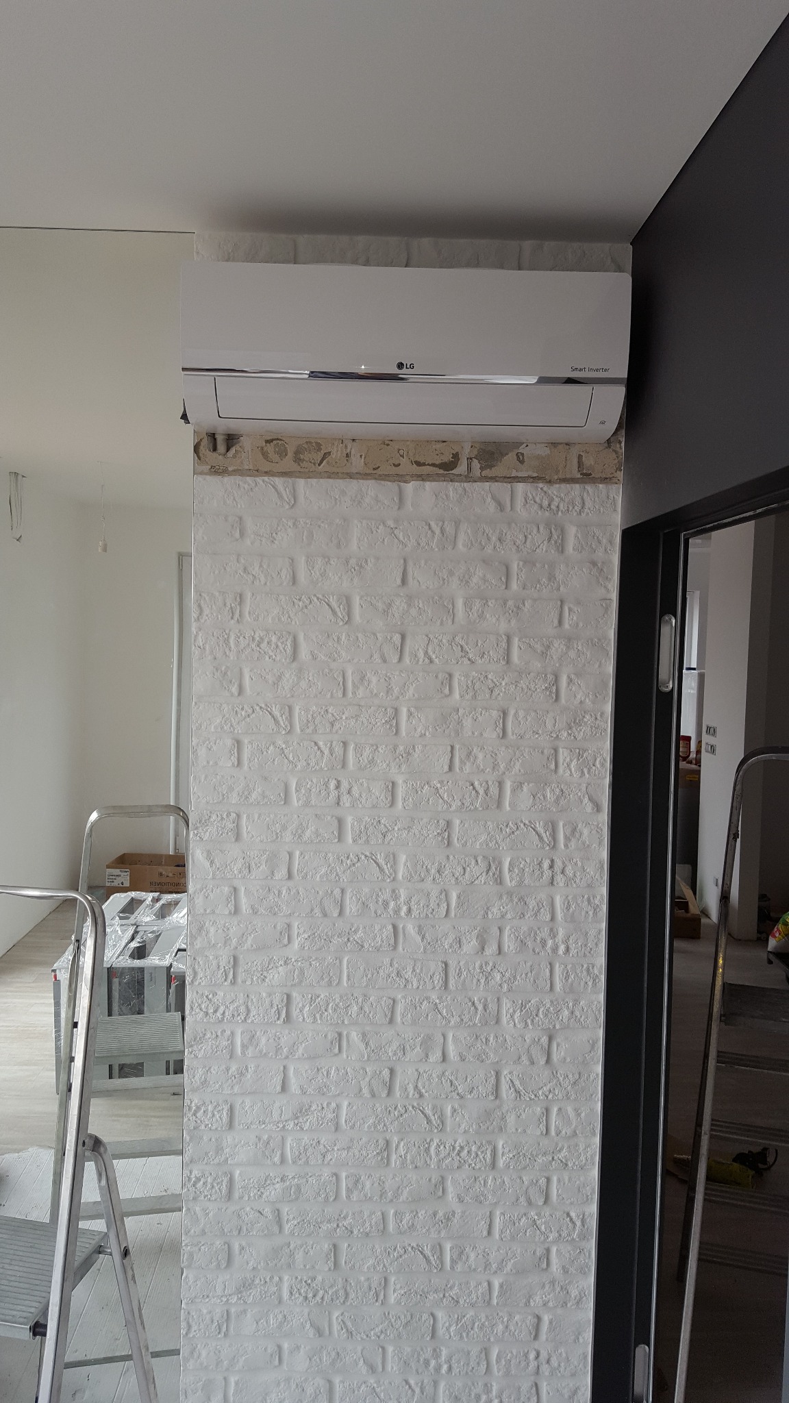 Zamontowany klimatyzator LG Smart Inverter na ścianie z białej cegły dekoracyjnej, widoczne drabiny malarskie i elementy wykończeniowe.