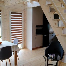 Logbar Sp. z o.o. - Jasne wnętrze domu z bali z drewnianymi schodami, oknami z roletami i nowoczesną kuchnią w tle.