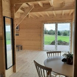 Logbar Sp. z o.o. - Jasne wnętrze domu z drewnianymi ścianami, podłogą i schodami, okrągły biały stół z krzesłami w części jadalnej, widok na zewnątrz przez duże okno.