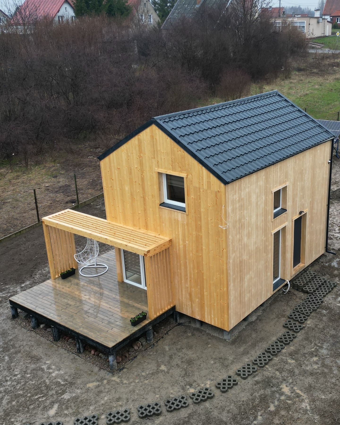 Nowoczesny, parterowy dom z jasnego drewna z czarnym dachem, tarasem z pergolą i wiszącym fotelem, widok z góry na posesję z ażurowymi płytami chodnikowymi.