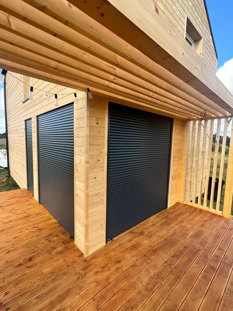 Drewniany budynek z czarnymi roletami zewnętrznymi, oświetlony wiszącymi żarówkami, z drewnianym tarasem i pergolą, widok z dołu.