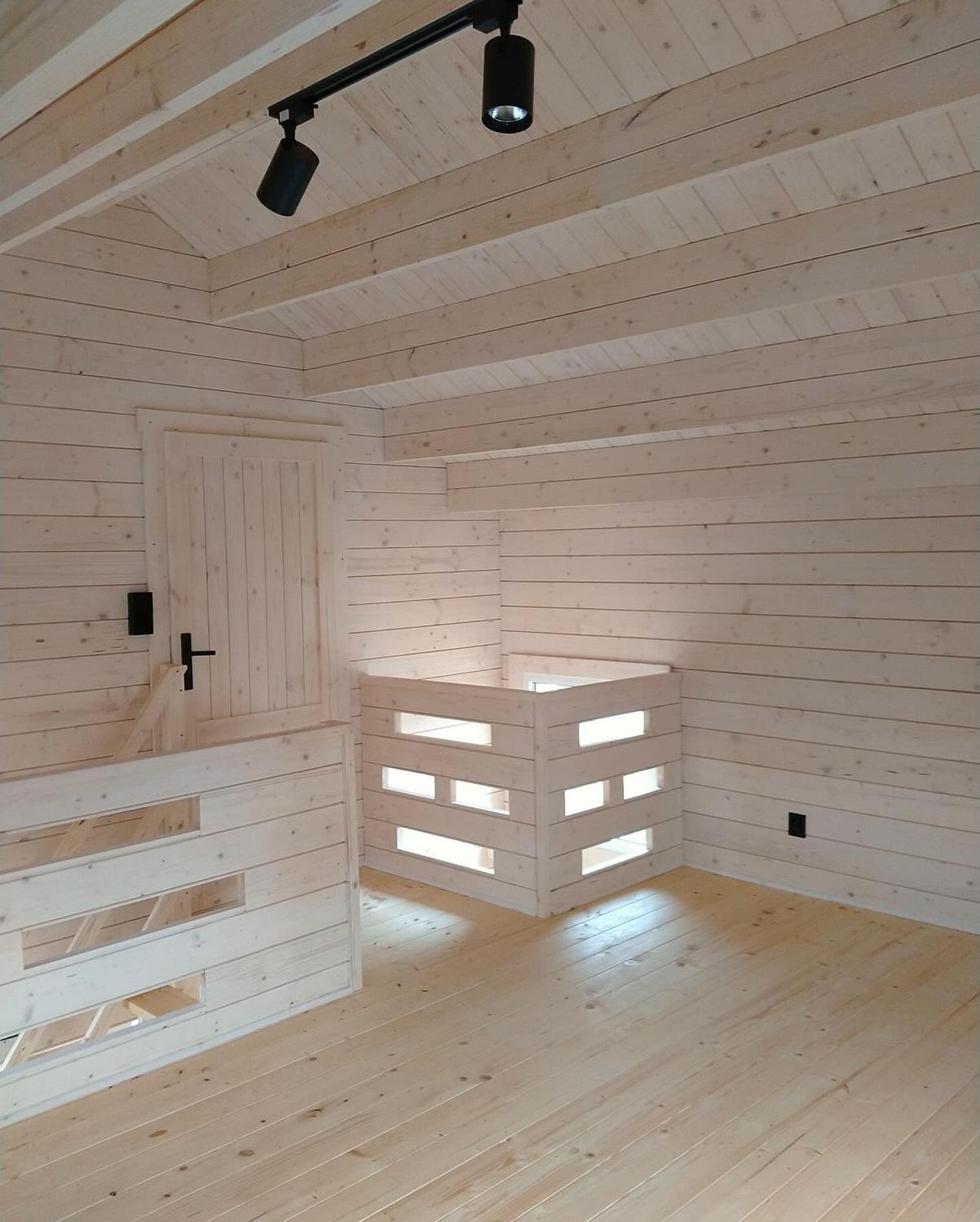 Wnętrze domu z bali, surowe drewno, widoczne belki stropowe, drewniana balustrada z prostokątnymi prześwitami, minimalistyczne oświetlenie punktowe.