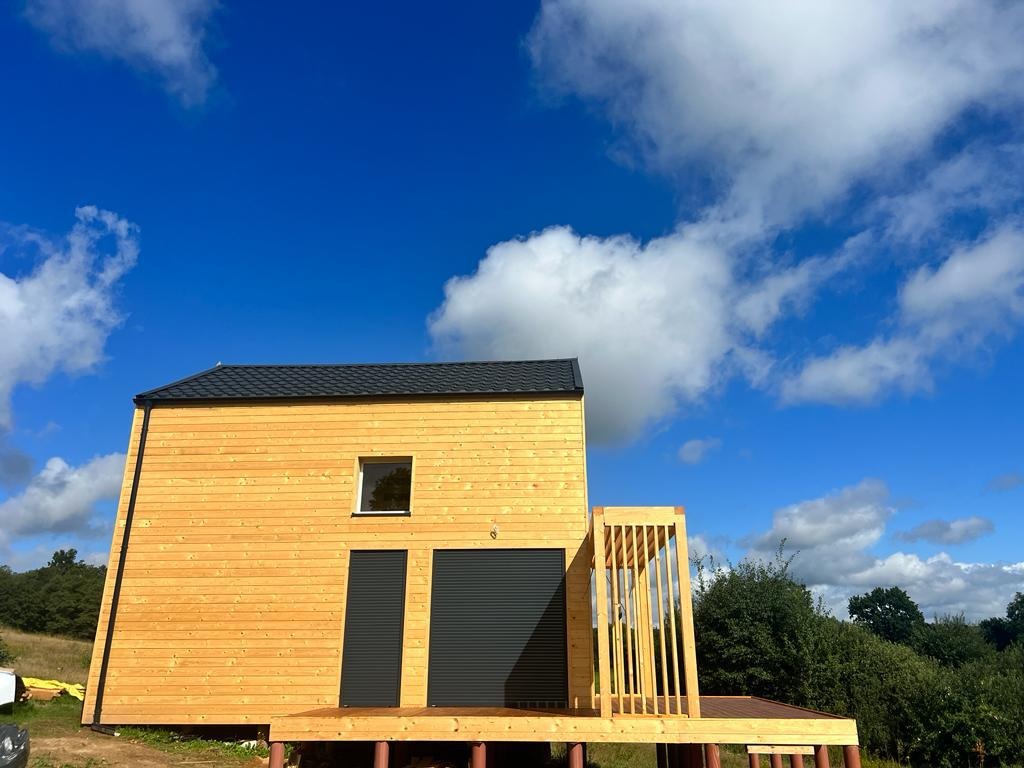 Nowoczesny dom o prostej bryle z jasną, drewnianą elewacją, ciemnym dachem i szarymi roletami, otoczony zielenią i błękitnym niebem z chmurami.