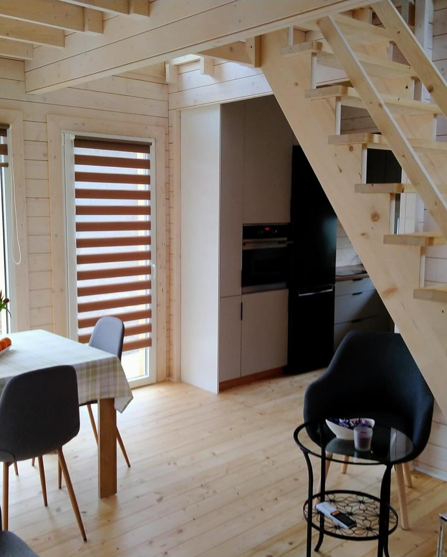 Jasne wnętrze domu z bali z drewnianymi schodami, oknami z roletami i nowoczesną kuchnią w tle.