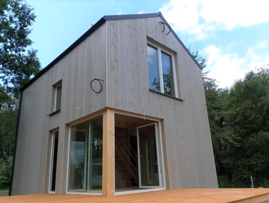 Nowoczesny dom z jasnego drewna z przeszkloną werandą i drewnianym tarasem, otoczony zielenią.