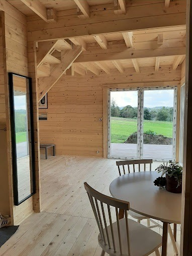 Jasne wnętrze domu z drewnianymi ścianami, podłogą i schodami, okrągły biały stół z krzesłami w części jadalnej, widok na zewnątrz przez duże okno.