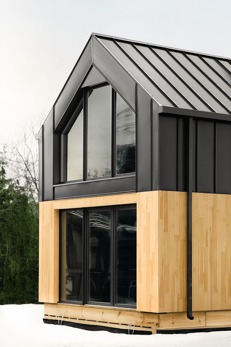 Fragment nowoczesnego domu z drewnianą elewacją i czarnym dachem z blachy na tle zimowego krajobrazu, widoczne duże okna.
