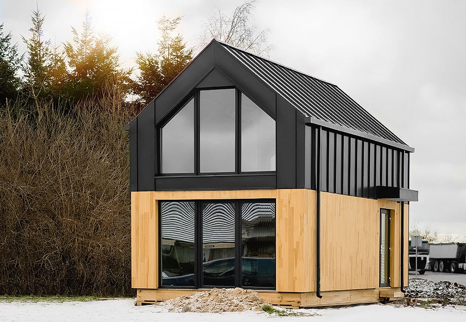 Dom M35 z czarną, metalową fasadą na górze i jasnym drewnem na dole, duże okna, widoczny fragment ciężarówki w tle, częściowo pokryty śniegiem.