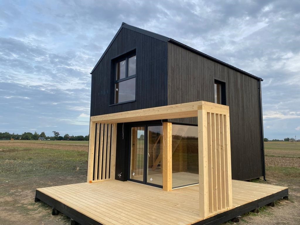 Czarny, dwukondygnacyjny dom szkieletowy z jasnym drewnianym tarasem i pergolą z pionowymi listwami, na tle pola i pochmurnego nieba.