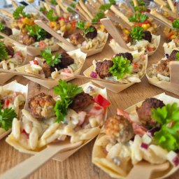 Catering & Cooking Corner - Mini porcje sałatki z klopsikami, podane w ekologicznych, drewnianych łódeczkach z dołączonymi mini łyżeczkami, udekorowane świeżą pietruszką. W tle widoczne inne przekąski.