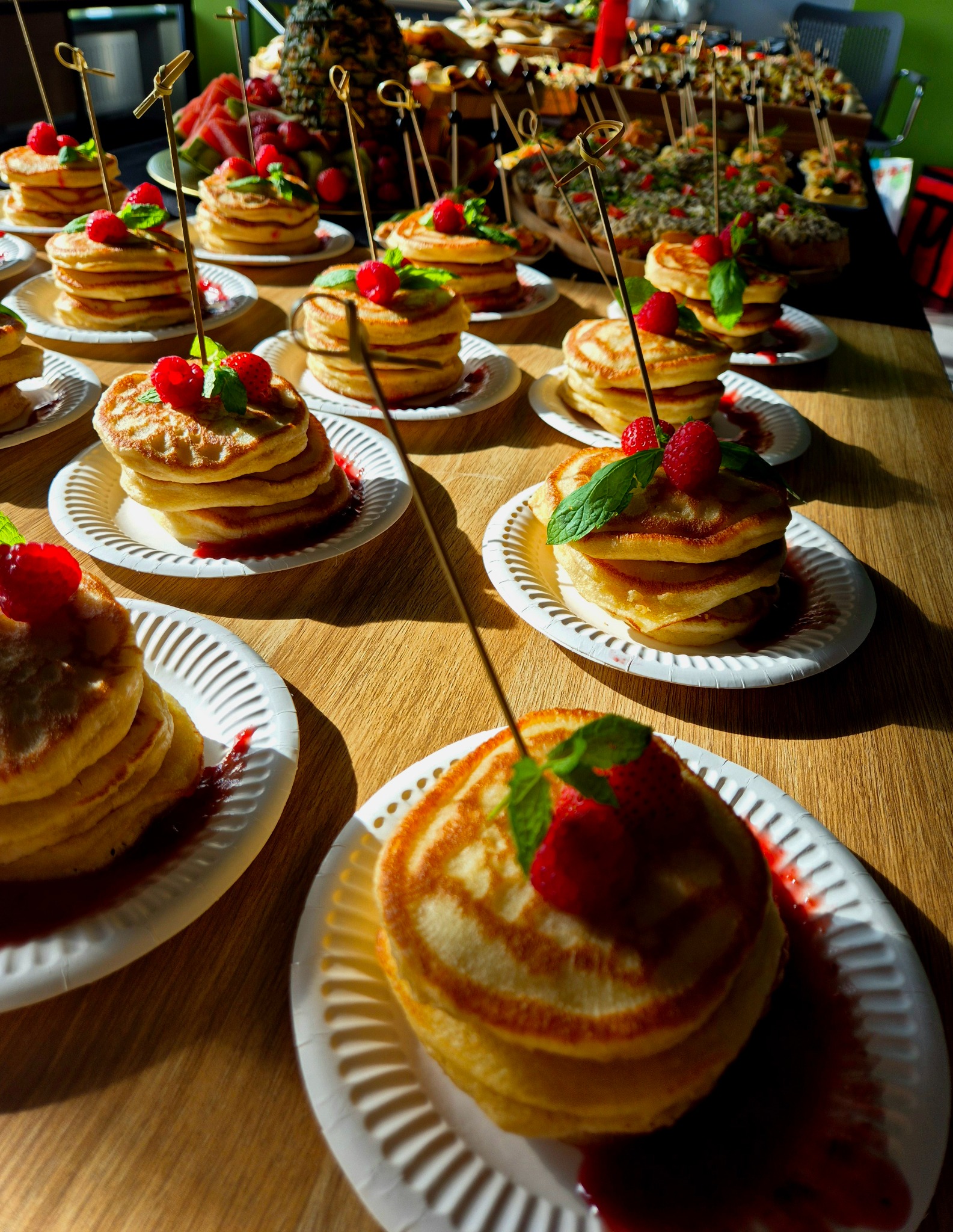Stosy puszystych naleśników na papierowych talerzykach, udekorowane malinami, listkami mięty i sosem owocowym, ustawione na drewnianym stole podczas cateringu, w tle widoczne inne przekąski...