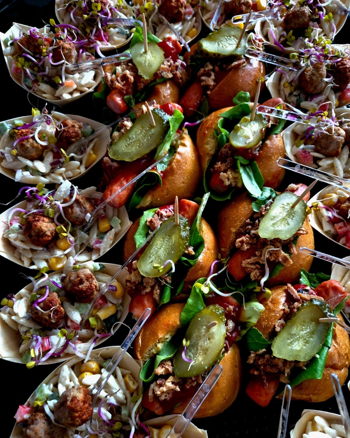 Widok z góry na aranżację finger food: miniaturowe hot dogi z sałatą, ogórkiem konserwowym i prażoną cebulką oraz sałatki makaronowe z pulpetami, podane w łódeczkach z jasnego drewna...