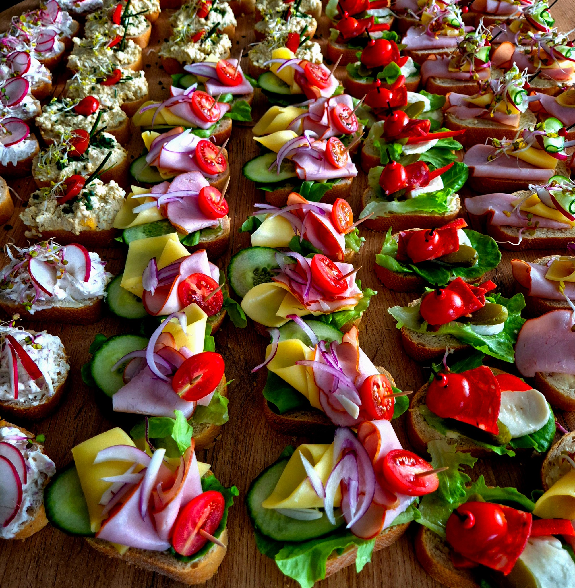 Różnorodne kanapki na kromkach chleba, udekorowane warzywami, wędlinami i pastami, ułożone rzędami na drewnianym blacie, niektóre spięte wykałaczkami.