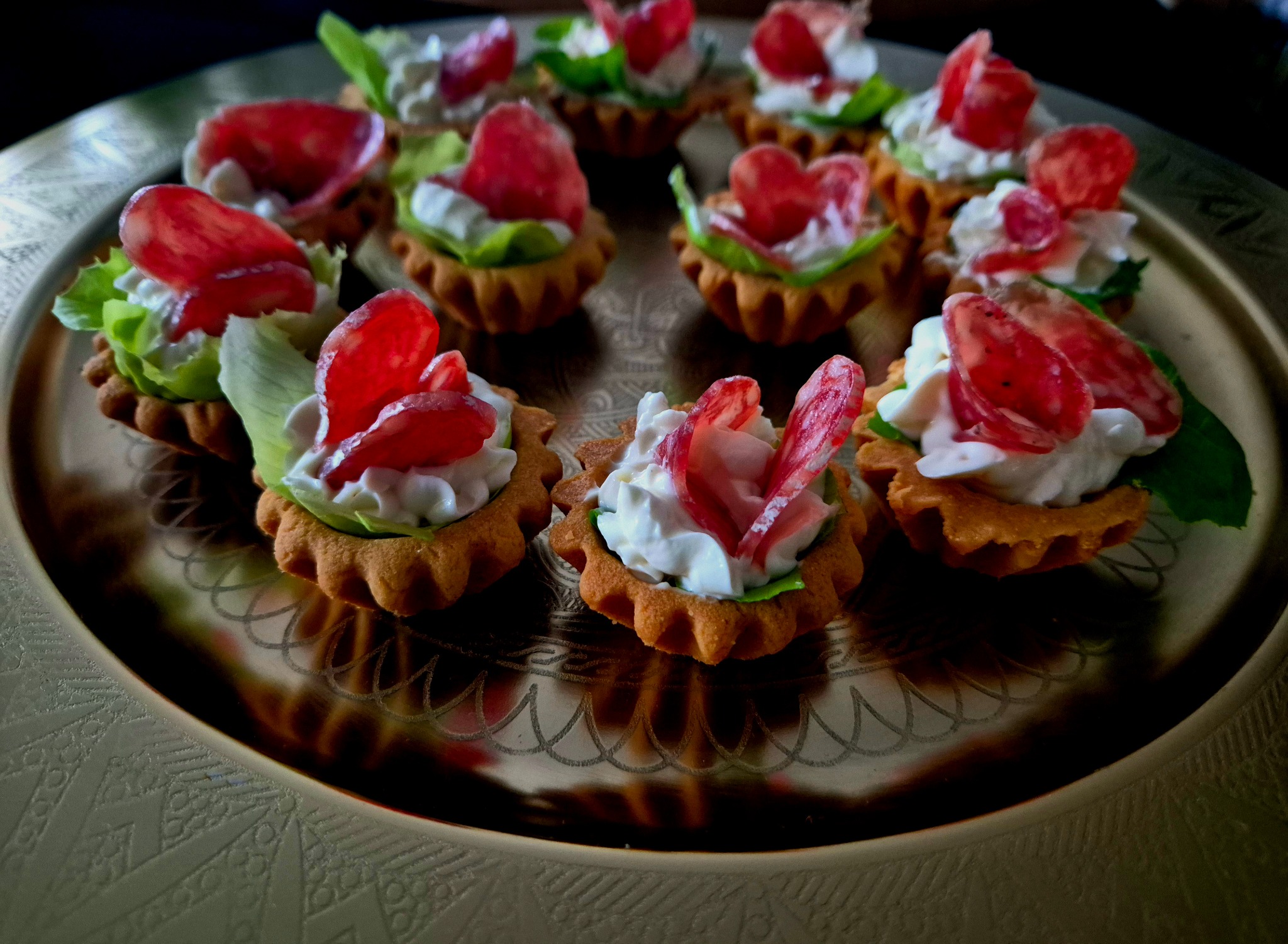 Talerz z tartaletkami: kruche babeczki wypełnione białym kremem, zieloną sałatą i plasterkami czerwonego salami, ułożone na złotej, dekoracyjnej tacy.