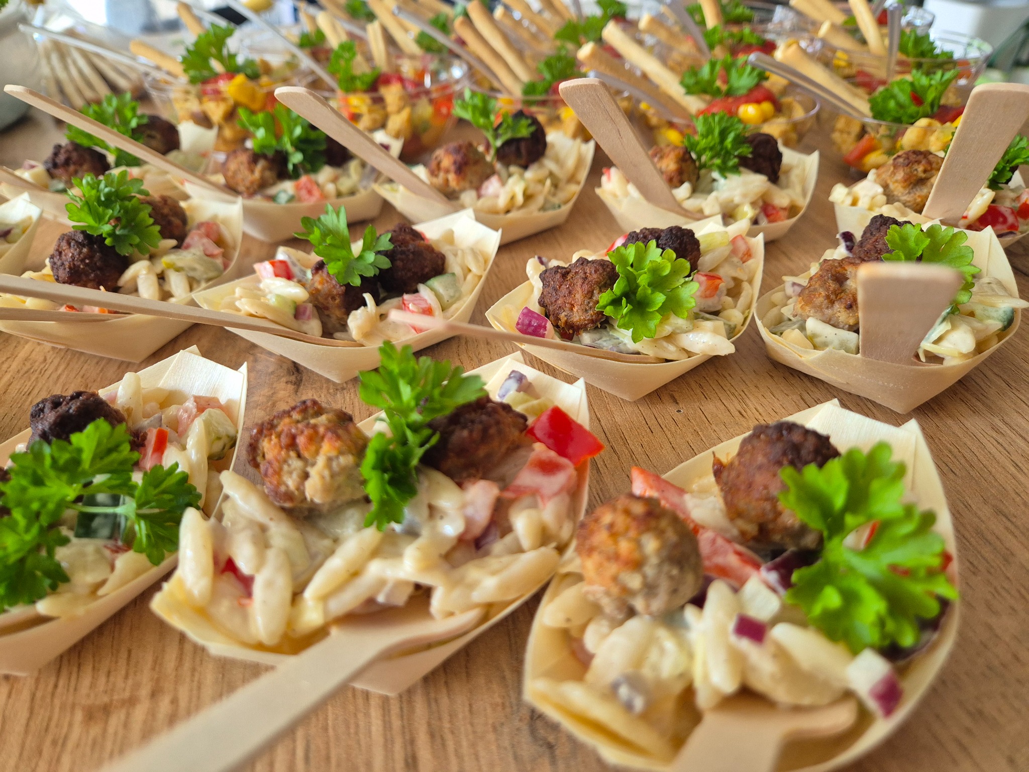 Mini porcje sałatki z klopsikami, podane w ekologicznych, drewnianych łódeczkach z dołączonymi mini łyżeczkami, udekorowane świeżą pietruszką. W tle widoczne inne przekąski.