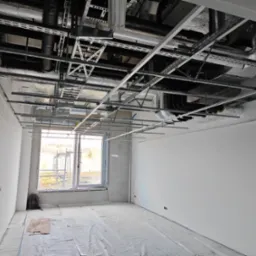 Wnętrze w trakcie remontu: widoczne metalowe profile sufitowe, instalacje wentylacyjne pod stropem, okno z widokiem na zewnątrz, podłoga zabezpieczona folią malarską, plecak w rogu pomieszczenia.